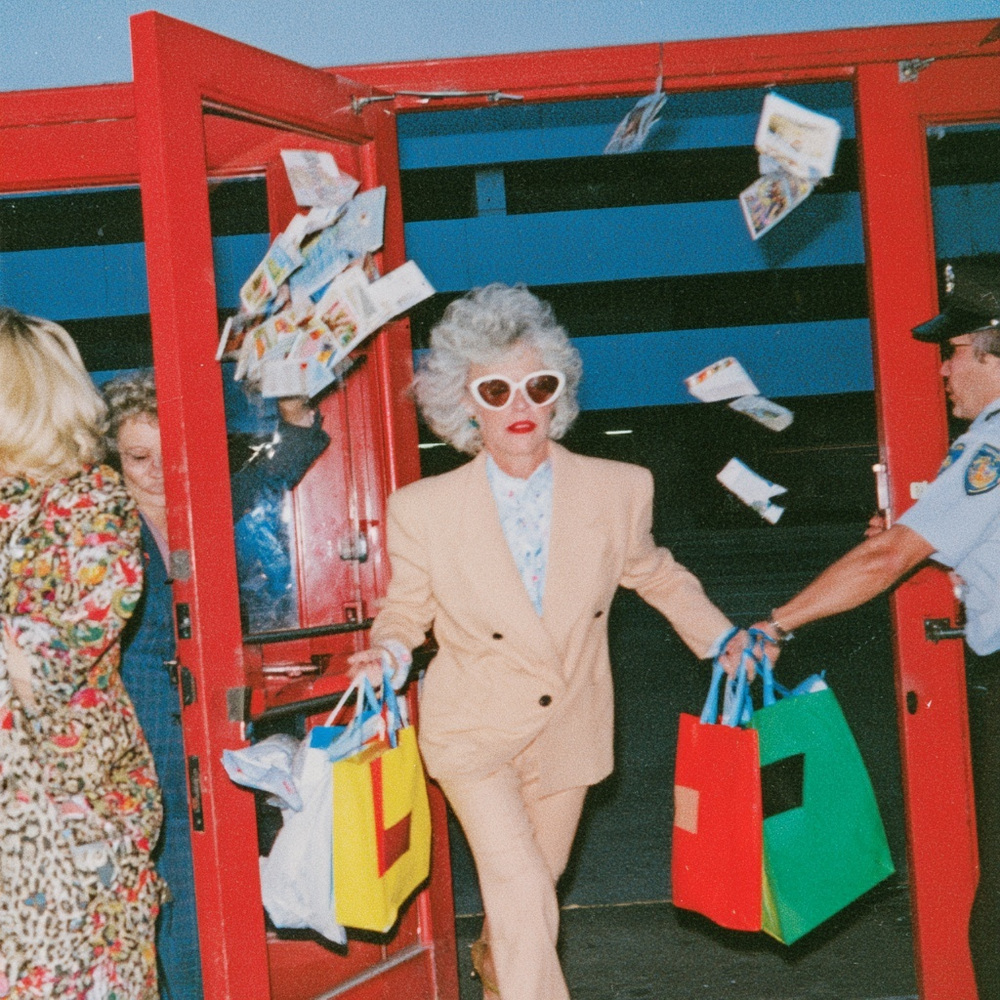 Silver-haired woman in a cream suit and sunglasses leaving red museum doors with bright shopping bags and flying postcards.