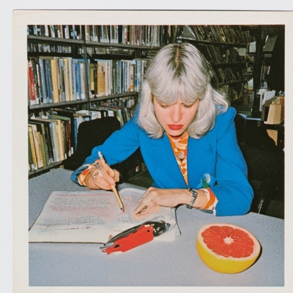 Silver-haired woman in a blue jacket writing at a library desk beside a red stapler and half a grapefruit.